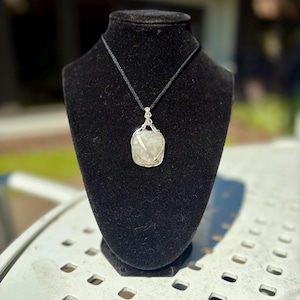 May include: A silver wire-wrapped pendant necklace with a light-colored stone, displayed on a black velvet bust. The pendant hangs from a black cord. The background is blurred, suggesting an outdoor setting.
