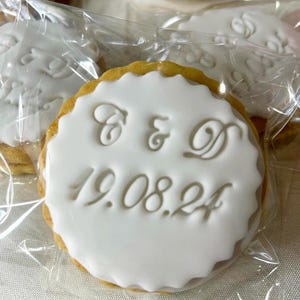 May include: A close-up shot of a decorated biscuit with white icing. The biscuit has a scalloped edge and the initials "C & D" and the date "19.08.24" are imprinted on the icing. The biscuit is packaged in clear cellophane.