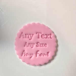 May include: A pink, circular stamp with a scalloped edge. The stamp is embossed with the words "Any Text, Any Size, Any Font" in a sans-serif font. The stamp is on a white surface.