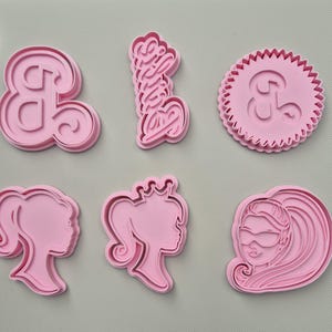 May include: A collection of pink plastic cookie cutters. The shapes include an ampersand, the word "Barbie" in script, and three female profile silhouettes. These cutters are suitable for baking and craft projects.