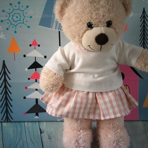 May include: A plush beige teddy bear with dark eyes and a brown nose. The bear is wearing a plain white short-sleeved shirt and a peach and white gingham patterned skirt with a gathered waistline, featuring a soft, fuzzy texture throughout.