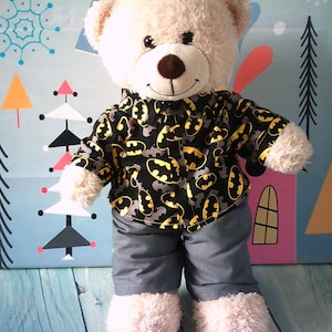 Può includere: Un orsacchiotto di peluche beige indossa una camicia nera abbottonata con un motivo a logo Batman giallo e nero e pantaloncini grigi. L'orso ha naso e bocca cuciti marrone scuro, occhi in plastica nera e si trova in posizione eretta.