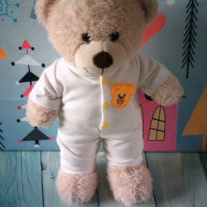 May include: A soft teddy bear dressed in a white onesie with a bear emblem. The bear has a brown nose and black eyes. The onesie has yellow button details. The background features a blue and teal pattern with geometric shapes.