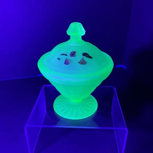 May include: A glowing green glass candy dish with a lid, set on a clear acrylic stand. The dish has a pedestal base and a decorative lid with painted accents. The glass appears to fluoresce under a black light.