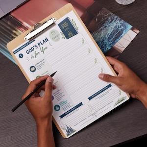 May include: A wooden clipboard holding a printed study sheet titled God's Plan is for You. The sheet features sections for notes, questions, and temple recommendations, decorated with small green leaf illustrations and blue accents, held by a metal clip.