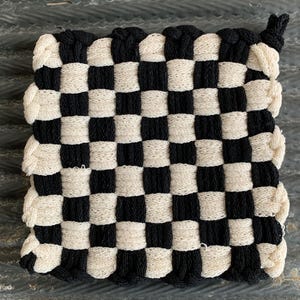 May include: A square, woven potholder with a checkerboard pattern of black and cream-colored squares. The potholder has a loop for hanging and is made of a thick, textured material. The design is simple and classic.