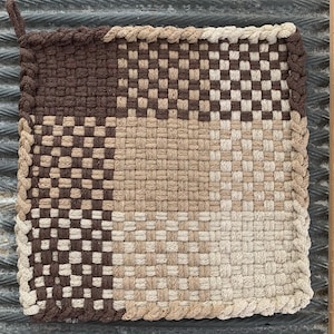 May include: A square, woven potholder in shades of brown and beige. The potholder is divided into four sections, each with a different pattern and color. A loop is attached to one corner for hanging.