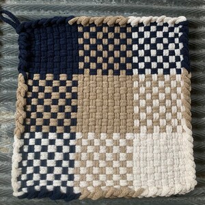 May include: Square woven pot holder with a loop for hanging. The design features a checkerboard pattern in navy blue, tan, and white. The edges are trimmed with a braided border in a matching tan color.
