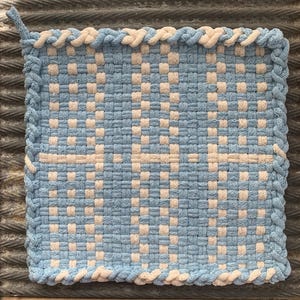 May include: A square, woven potholder in light blue and cream. The potholder features a checkerboard pattern with a cream-colored border and a loop for hanging. The design is symmetrical, with a horizontal cream stripe across the center.