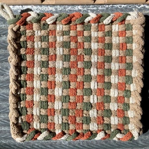 May include: A square woven pot holder with a braided border. The pot holder is made of natural fibers in shades of brown, green, and orange.