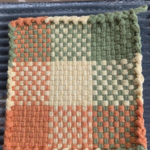 May include: A woven pot holder with a checkered pattern in shades of green, orange, and yellow. The pot holder has a braided border.
