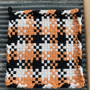 May include: A woven pot holder with a black, white, and orange checkered pattern. The pot holder has a fringed edge.