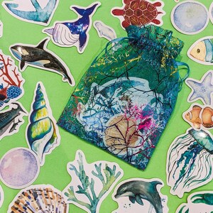 Peut inclure: Une collection d'autocollants color&eacute;s sur le th&egrave;me de l'oc&eacute;an repr&eacute;sentant une vari&eacute;t&eacute; de cr&eacute;atures marines, notamment une baleine, une tortue, une m&eacute;duse, une &eacute;toile de mer, un coquillage et un r&eacute;cif corallien. Les autocollants sont dispos&eacute;s sur un fond vert.