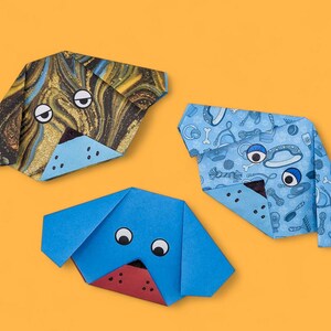 Pode incluir: Tr&ecirc;s c&atilde;es de papel origami. Um c&atilde;o &eacute; azul com olhos pretos, outro &eacute; azul com um padr&atilde;o de osso de c&atilde;o e o &uacute;ltimo &eacute; marrom e dourado com olhos pretos.