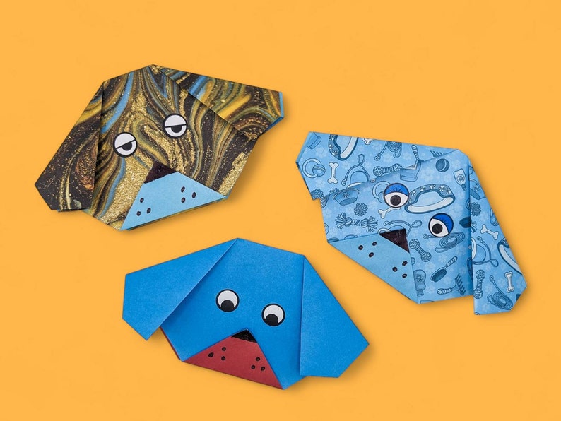 Pode incluir: Tr&ecirc;s c&atilde;es de papel origami. Um c&atilde;o &eacute; azul com olhos pretos, outro &eacute; azul com um padr&atilde;o de osso de c&atilde;o e o &uacute;ltimo &eacute; marrom e dourado com olhos pretos.