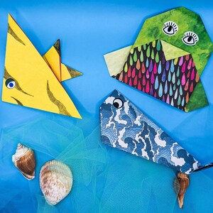 Peut inclure: Trois animaux en papier origami, un poisson jaune, un poisson bleu et un animal en forme de nuage vert et noir, sont dispos&eacute;s sur un fond bleu avec un tissu en filet bleu et blanc en dessous.
