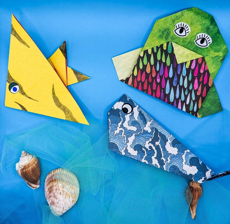 Peut inclure: Trois animaux en papier origami, un poisson jaune, un poisson bleu et un animal en forme de nuage vert et noir, sont dispos&eacute;s sur un fond bleu avec un tissu en filet bleu et blanc en dessous.