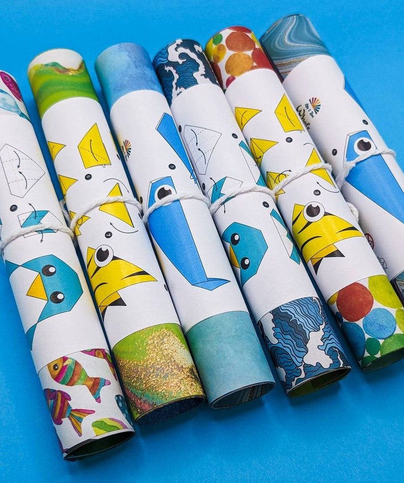 Peut inclure: Un ensemble de huit instructions d'origami en papier enroul&eacute;es, avec des motifs et des illustrations de poissons color&eacute;s. Les instructions sont attach&eacute;es avec une ficelle blanche et ont un fond bleu.