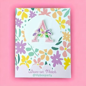 May include: Pink iron-on patch featuring a floral design in the shape of the letter A. The patch is displayed on a white card with a floral pattern and the text "Iron-on Patch @lilybeeparty".