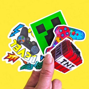 May include: A set of five colourful vinyl stickers featuring video game and pixel art themes. The stickers include a green pixelated block, a red and black video game controller, a red and blue pixelated character, and a red and white TNT block with the word "TNT" printed on it. The stickers are perfect for decorating laptops, water bottles, notebooks, and other surfaces.