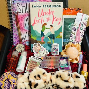 Pode incluir: Uma caixa de presente cheia de vários itens, incluindo um livro de Lana Ferguson intitulado "Under Loch and Key", um caderno, uma caneta, adesivos, uma faixa de cabelo, produtos de banho e café. A caixa é decorada com papel vermelho e tem uma variedade de cores.
