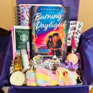 Pode incluir: Uma caixa de presente cheia de um livro intitulado "Burning Daylight", um caderno, chocolate quente Starbucks, produtos de banho, uma vela e uma faixa de cabeça colorida. A caixa é forrada com papel de seda roxo.
