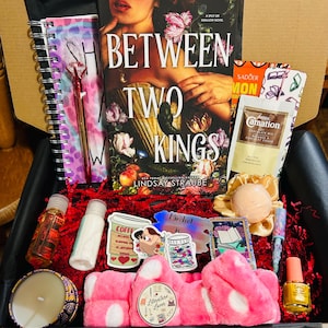 Pode incluir: Uma caixa de presente cheia de um livro intitulado "Between Two Kings" de Lindsay Straube, uma faixa de cabelo rosa, adesivos, um caderno, uma caneta, uma vela, uma bomba de banho, esmalte de unhas e outros itens. A caixa é preenchida com papel vermelho.