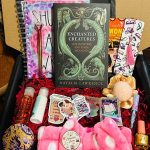 Pode incluir: Uma caixa de presente cheia de vários itens, incluindo um livro intitulado "Enchanted Creatures", um caderno espiral, uma caneta, marcadores, adesivos, uma bomba de banho, verniz de unhas e uma faixa de cabeça rosa Literature Lover. A caixa é forrada com papel vermelho.