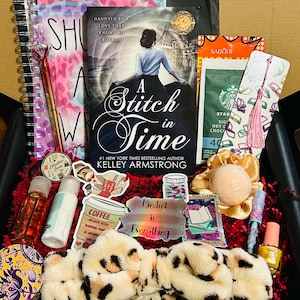 Pode incluir: Uma caixa de presente cheia de vários itens, incluindo um livro intitulado "A Stitch in Time" de Kelley Armstrong, um caderno espiral, uma caneta, adesivos e produtos de beleza. A caixa contém enchimento de papel vermelho e uma faixa de cabeça com estampa de leopardo.