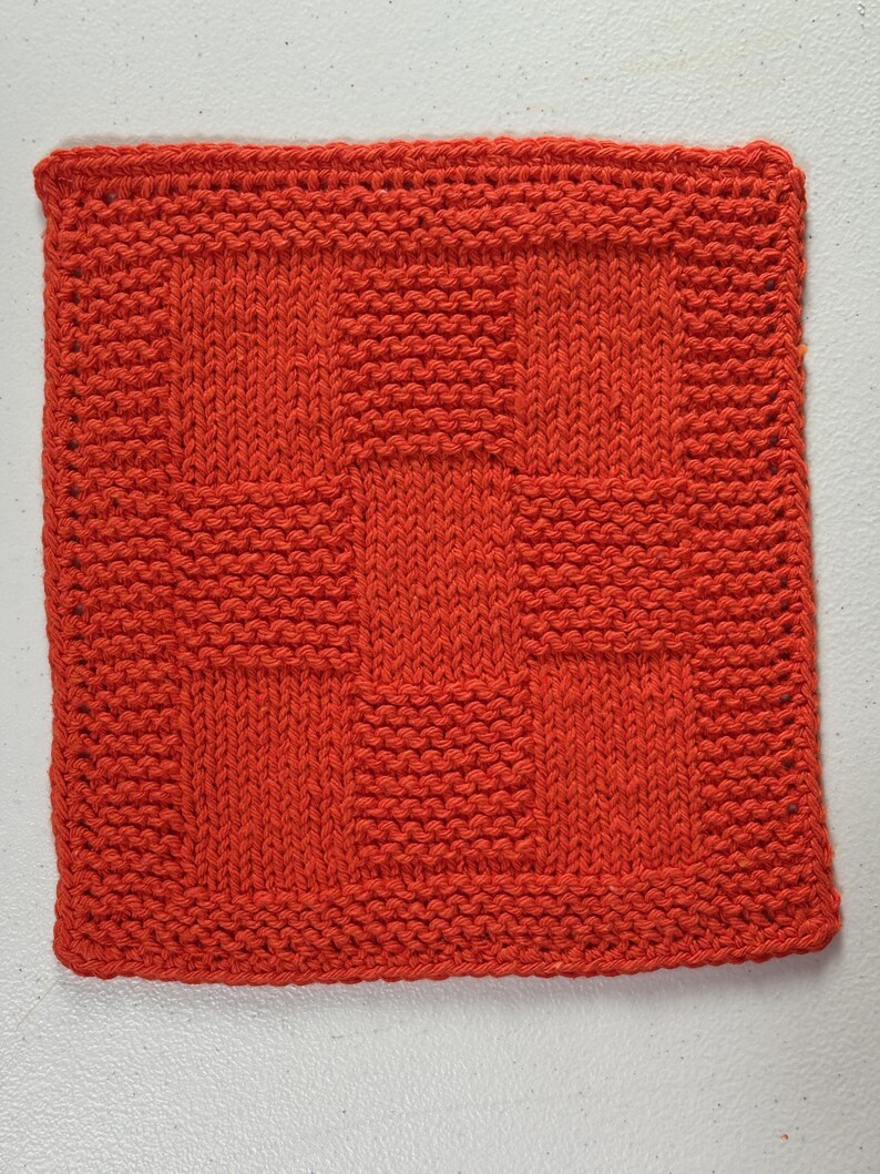 May include: A square, hand-knitted washcloth in a vibrant orange color. The textile features a textured pattern created by alternating blocks of stockinette stitch and garter stitch, forming a grid-like design across the entire surface.