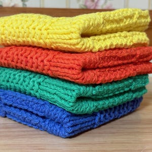 May include: A stack of four folded square crochet cloths in bright solid colors. The top cloth is yellow, followed by an orange cloth, a green cloth, and a blue cloth at the bottom. Each piece features a consistent textured knit pattern.