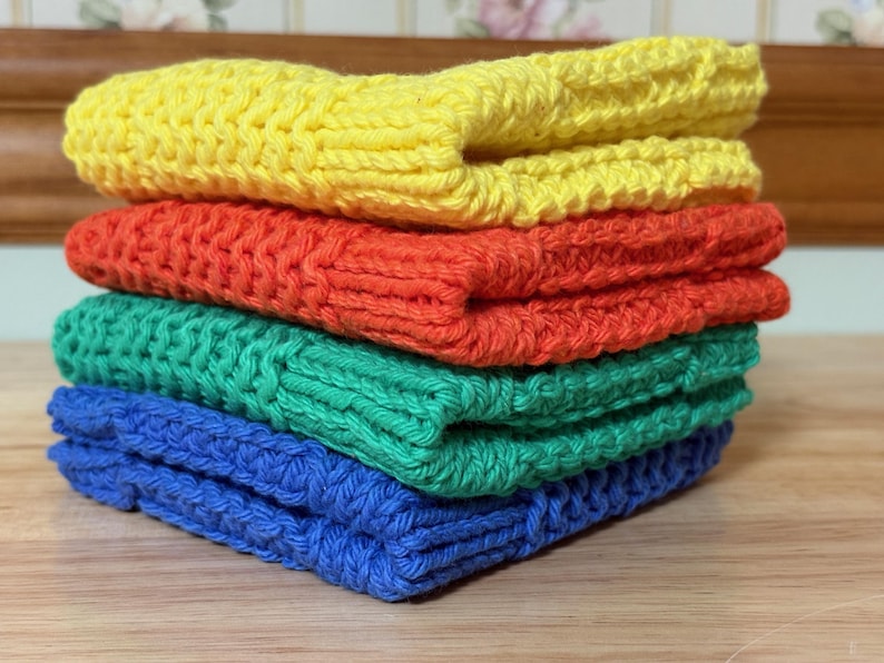 May include: A stack of four folded square crochet cloths in bright solid colors. The top cloth is yellow, followed by an orange cloth, a green cloth, and a blue cloth at the bottom. Each piece features a consistent textured knit pattern.
