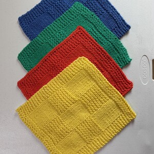 May include: A set of four square knitted dishcloths arranged in a stack. The cloths are solid blue, green, red, and yellow, each featuring a textured basketweave pattern with a garter stitch border. The items are displayed in a layered formation.