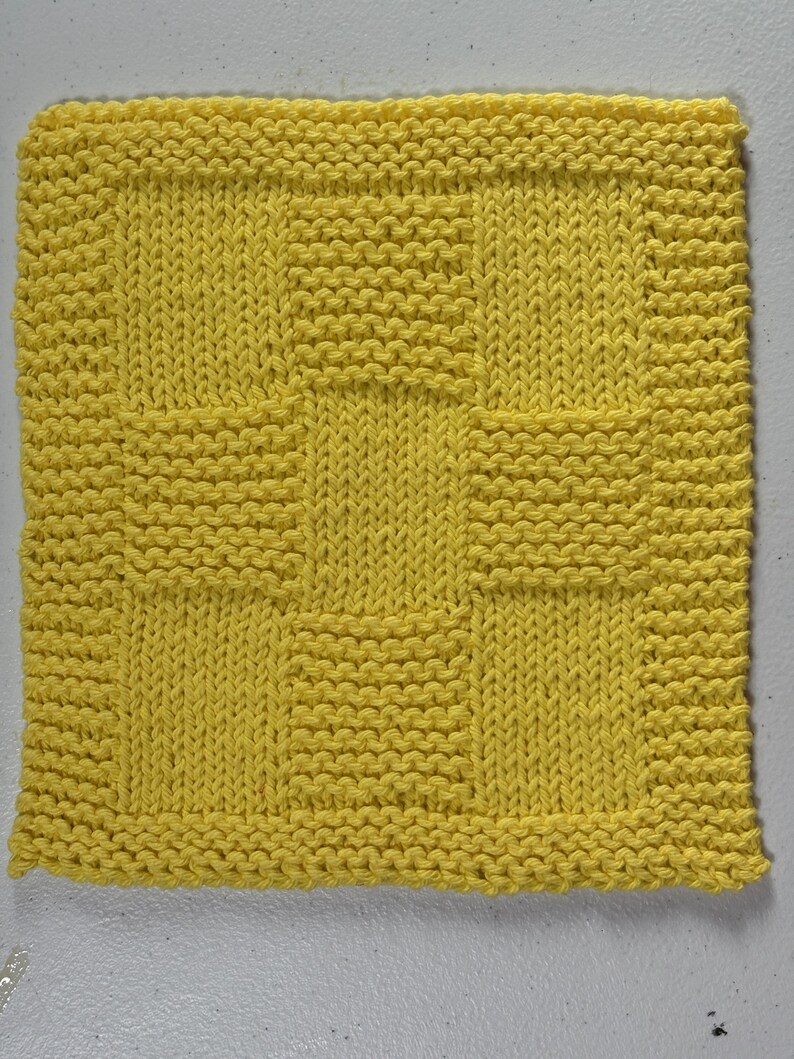 May include: A square, bright yellow knitted dishcloth featuring a textured pattern of alternating garter stitch and stockinette stitch blocks. The design consists of a central square surrounded by a border of smaller rectangular blocks in a grid.