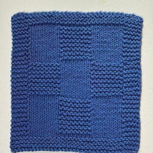 May include: A square blue knitted washcloth featuring a checkerboard pattern. The design alternates between sections of garter stitch and stockinette stitch, creating a textured grid effect across the entire surface of the blue fabric square.