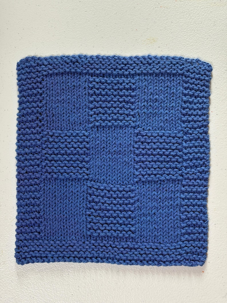 May include: A square blue knitted washcloth featuring a checkerboard pattern. The design alternates between sections of garter stitch and stockinette stitch, creating a textured grid effect across the entire surface of the blue fabric square.