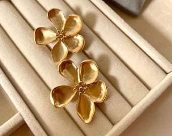 May include: A pair of gold-colored flower-shaped earrings. Each earring features five petals and a textured center, resting on a beige jewelry display. The earrings have a shiny, metallic finish, reflecting light.