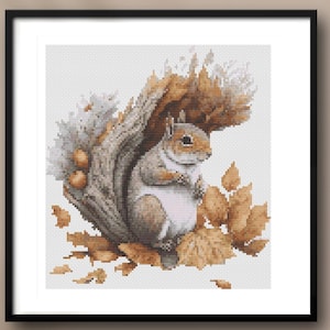May include: A framed cross-stitch artwork depicting a squirrel in a tree trunk, surrounded by autumn leaves. The squirrel is in shades of grey and white, with brown leaves and a light background. The artwork is presented in a black frame.