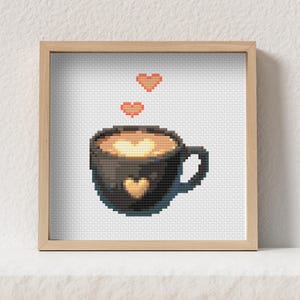 Può includere: Un'opera d'arte a punto croce incorniciata con una tazza di caffè pixelata con un design a cuore. La tazza nera ha un cuore dorato ed è riempita di liquido marrone chiaro. Due cuori arancioni fluttuano sopra la tazza, su sfondo bianco.