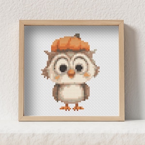 May include: A framed cross-stitch picture of a cartoon owl wearing a pumpkin hat. The owl has large, round eyes, a light brown body, and orange and brown accents. The artwork is set against a white background.