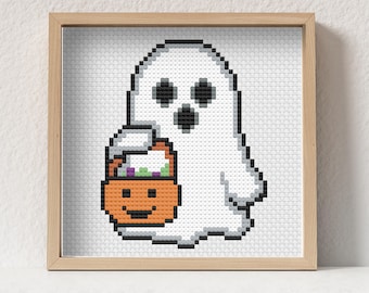 Puede incluir: Un punto de cruz enmarcado con un fantasma blanco pixelado sosteniendo un cubo de caramelos de calabaza naranja. El fantasma tiene ojos y boca negros. El cubo de caramelos tiene una cara sonriente y está lleno de caramelos morados y verdes. El marco es de madera clara.