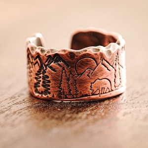 May include: A copper ring with a textured surface and an engraved design of mountains, trees, and a bear. The ring is a wide band with a rustic look.