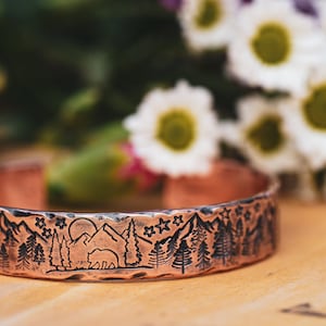 May include: A copper bracelet with a hammered texture and an engraved design of a mountain range, a bear, and stars.