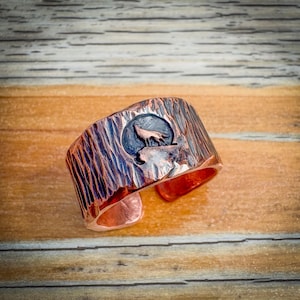 May include: A copper ring with a textured surface and a wolf silhouette design. The wolf is howling at the moon.