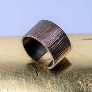 May include: A wide copper band ring with a textured hammered finish. The ring is made of metal and has a rustic look.