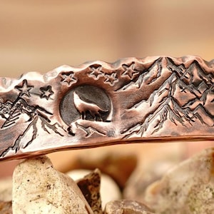 May include: A copper-colored cuff bracelet with a mountain and wolf design. The bracelet features a detailed landscape with mountains, trees, stars, and a howling wolf in a circular moon. The cuff is resting on a bed of rocks.
