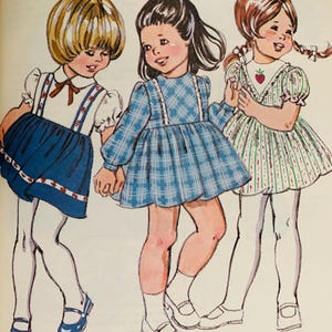 May include: Three girls wearing dresses. The girl on the left is wearing a blue and white dress with a white collar and a red bow. The girl in the middle is wearing a blue plaid dress with a white collar. The girl on the right is wearing a green and white striped dress with a red strawberry on the front.