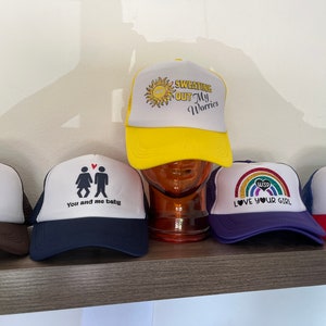 Peut inclure: Cinq casquettes de camionneur différentes avec des designs variés. Les casquettes sont blanches avec un dos en mesh et des visières de différentes couleurs. Les motifs comprennent un soleil avec le texte "Sweating Out My Worries", un couple avec le texte "You and me baby", un arc-en-ciel avec le texte "Love Your Girl", un logo de l'État du Colorado avec le texte "June 21", et un Bigfoot avec des lunettes de soleil.