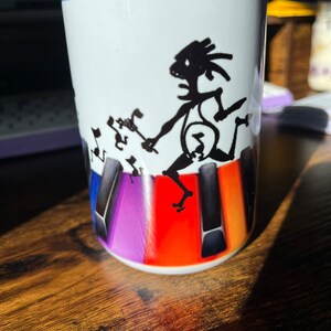 K&ouml;nnte beinhalten: Wei&szlig;e Keramiktasse mit blauem Rand, die eine schwarze Silhouette einer Person zeigt, die ein Keyboard mit Noten spielt. Die Tastatur ist mit bunten Tasten in den Farben Lila, Orange und Rot dargestellt.