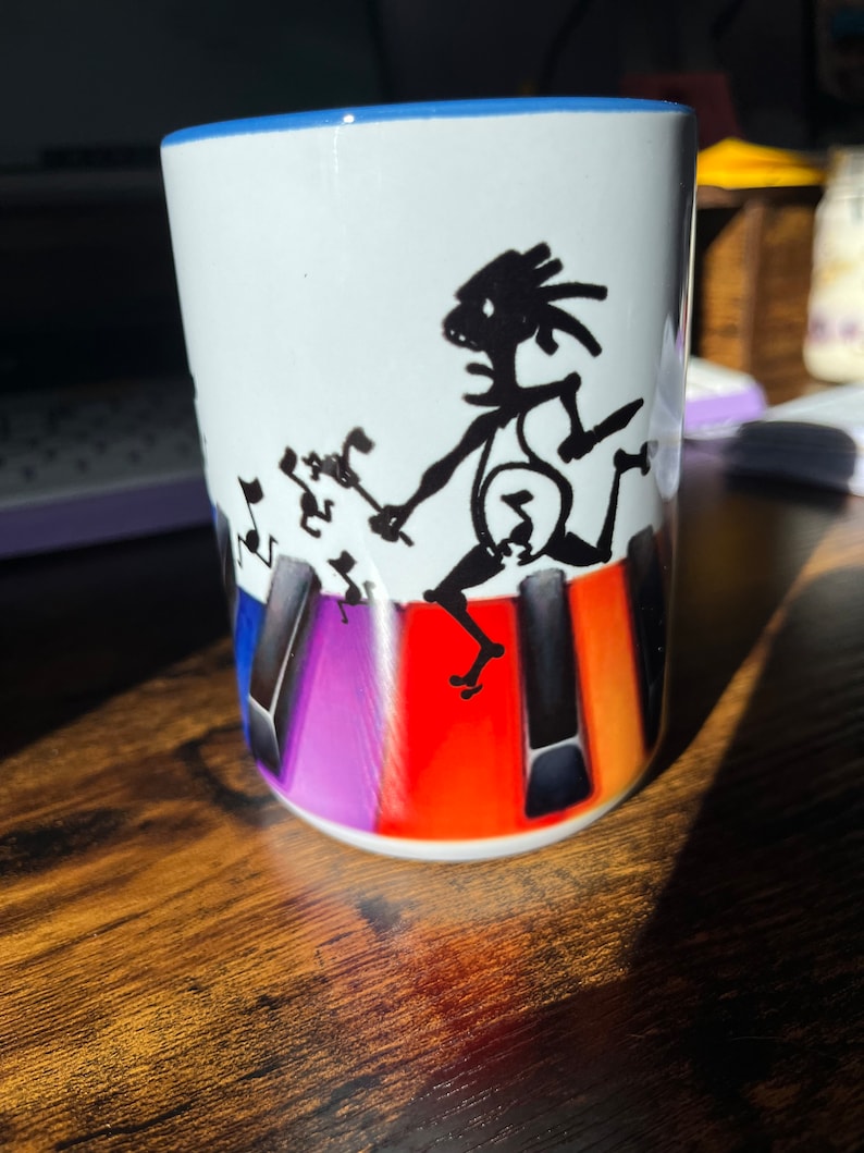 K&ouml;nnte beinhalten: Wei&szlig;e Keramiktasse mit blauem Rand, die eine schwarze Silhouette einer Person zeigt, die ein Keyboard mit Noten spielt. Die Tastatur ist mit bunten Tasten in den Farben Lila, Orange und Rot dargestellt.